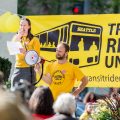 Wilson holds a bullhorn and speaks at a pre-pandemic rally standing in from or a big yellow Transit Riders Union banner.