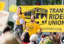 Op-Ed: Looking Back at the Movement That Elected Katie Wilson Wilson holds a bullhorn and speaks at a pre-pandemic rally standing in from or a big yellow Transit Riders Union banner.