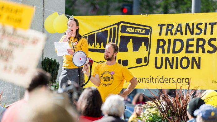 Katie-Wilson-Transit-Riders-Union-Seattle Wilson holds a bullhorn and speaks at a pre-pandemic rally standing in from or a big yellow Transit Riders Union banner.