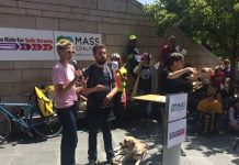 Monthly Meetup This Tuesday Featuring the MASS Coalition Anna Zivarts hold the microphone at a podium next to a bearded man with a guide dog with a crowd of people in bicycle helmets behind. Signs read "MASS Coalition" and "Ride for Safe Streets"