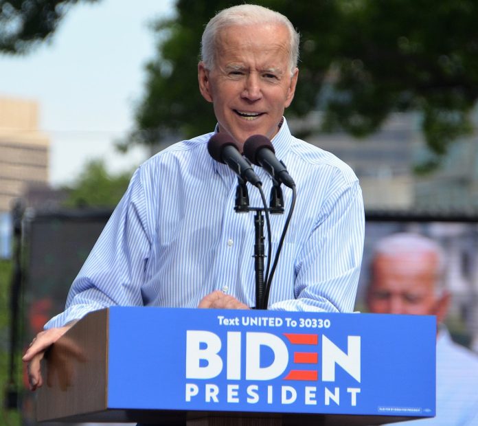 1212px-Joe_Biden_kickoff_rally_May_2019 Joe Biden in a blue collar behind a lectern with Biden for President on it.