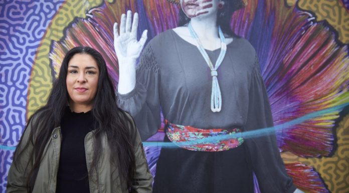 Colleen Echohawk campiagn photo with mural Colleen Echohawk with a colorful mural in the background showing an indigenous with a handprint over her lips.