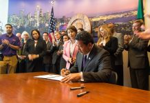 Centrists Turn to Old Formula: Crime Fears and More Money for Cops Bruce Harrell sits at a desk signing some orders at City Hall with a dozen supporters looking on.