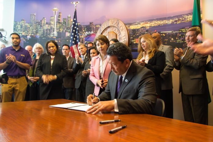 bruce harrell signs orders during his brief mayor stint Bruce Harrell sits at a desk signing some orders at City Hall with a dozen supporters looking on.