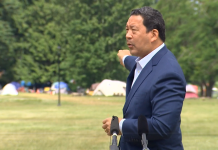 Durkan and Harrell Pressure Seattle Public Schools to Remove Homeless Encampments Harrell wears a blazer and gestures toward tents in the background.