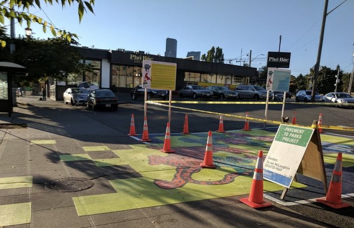 A photograph of a parking lot that has been painted with a brightly colored mural. Traffic cones mark the area surrounding part of the mural, and an A frame sign reads "pavement to parks."