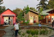 Op-Ed: How Mobile Villages Could Tackle Seattle’s Homelessness Emergency Tiny houses are oriented around a central green space where four people are standing. A couple is standing on the doorstep of a home in the background.