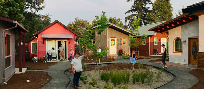 Tiny houses are oriented around a central green space where four people are standing. A couple is standing on the doorstep of a home in the background.