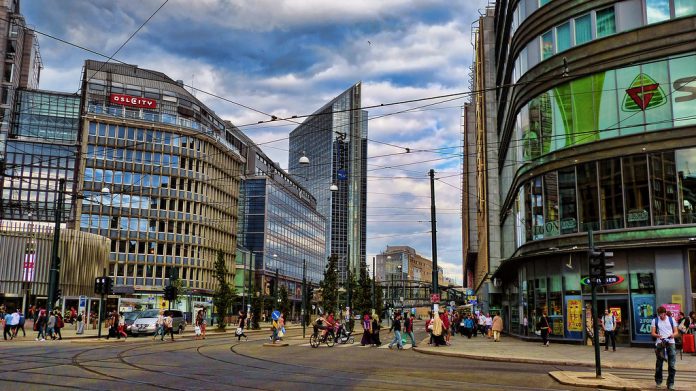 Oslo City by Miradortigre A photo of a city street without cars. There are tram lines and people walking and biking.