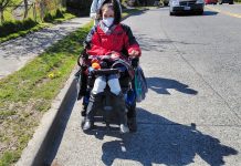 Week Without Driving, a Challenge with a Purpose A woman wearing a red jacket and face mask rides her power wheelchair in the road with a person walking behind her and a car approaching.