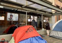 Op-Ed: Harrell’s Record on Homelessness Is Cynical, Cruel, and Cosmetic A photo of a tent in front of a glass window with exhibiting large portrait style photos of people.