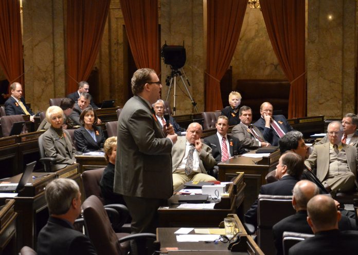 Chair of the Senate Transportation Committee Marko Liias A photo of a man wearing a suit in a senate chamber standing and speaking while other people in suits sit and listen.