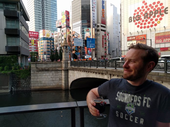 IMG_20160522_174732 Owen Pickford holding a beer, wearing a Sounders shirt in front of a bridge, river and large towers in Tokyo.