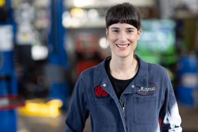 Marie_Perez_09_2022_1600 A young dark-haired woman in her blue autobody shop overalls.
