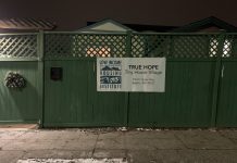 Tiny House Villages Take on Freezing Temperatures A sign for True Hope Tiny House Village on a green fence in front of a snowy sidewalk.