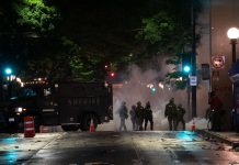 OPA, Seattle Police Violated Medical Privacy of Protester, Unearthed Investigation Reveals In a night time image, dark police officer silhouettes emerge from an armored truck amidst a backdrop of a cloud of tear gas.