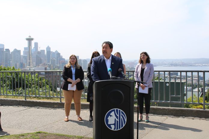 Bruce-Harrell-Press-Conference Bruce Harrell in a suit standing at a lectern. MLK Labor’s Executive Secretary-Treasurer Katie Garrow is to the left and OSE Director Jessyn Farrell to the right. A hazy Seattle skyline is in the background from Queen Anne vantage.