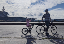 Bremerton’s Bike Network Approaches Its Moment of Truth Man on a bicycle with kiddycrank tandem attachment his daughter in pink dress is riding. Behind them is the USS Kitty Hawk aircraft carrier at Sinclair Inlet in Bremerton.