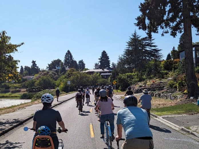 A procession of bicyclists as far as the eye can see down the middle of Lake Washington Boulevard with lots of trees along the shore.