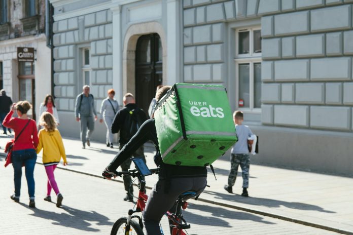 kai-pilger-dwBZLRPhHjc-unsplash A bike delivery person has a large green box with Uber Eats logo on it strapped to their back while riding down an urban streets with lots of pedestrians.