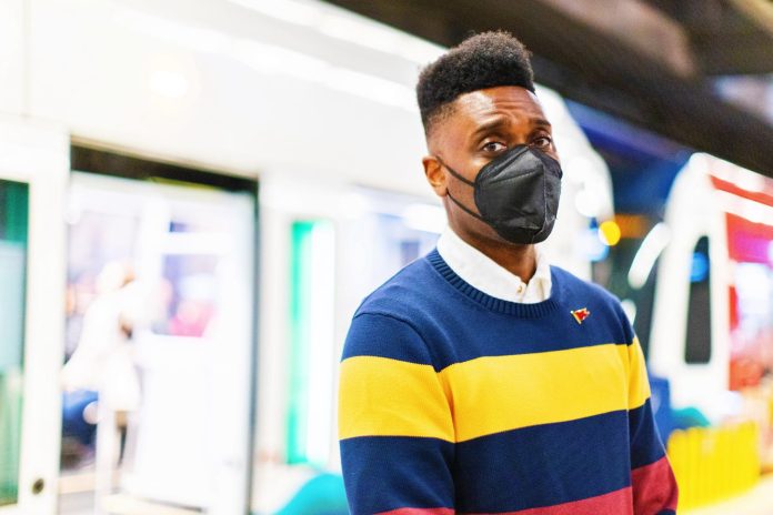 Shaun Scott is a Black man with a hi-top fade and wears an N95 mask while standing in front of a Link light rail train on a station platform.