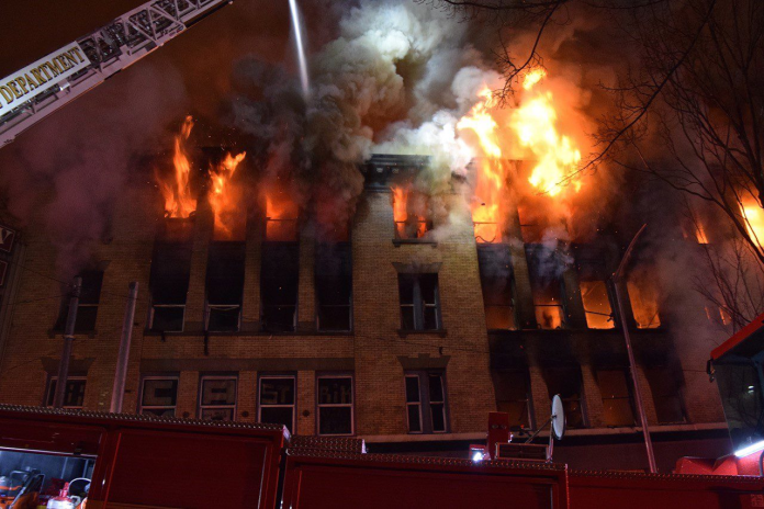 image-36 Fire and smoke pours out of upper story windows of an old brick building with a fire crane seeking to control them.