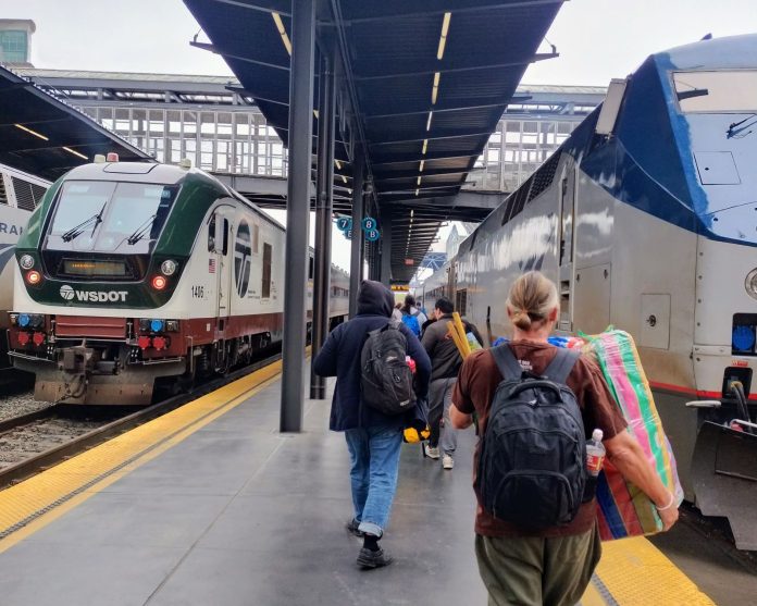 Amtrak passengers at Station by Ryan Packer Passengers lug their suitcases toward the train under an awning