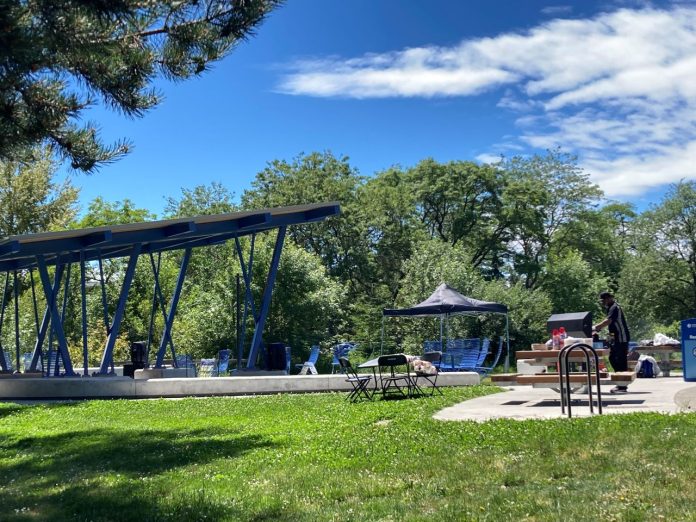 A large shelter provides rain protection over the stage alongside picnic table and playground equipment and a lawn with a row of trees in the background.