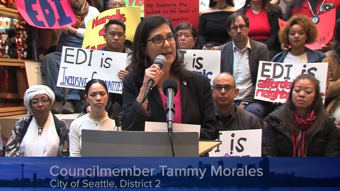 Tammy-Morales-at-EDI-Coalition-press-conference-by-Seattle-Channel Morales speaks at a podium and several rows of supporters hold signs in support of the EDI program and against the budget raid.