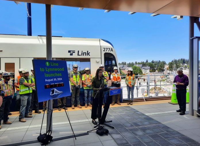 Terri-Mestas-at-Lynnwood-City-Center-Trumm-20240607 Mestas stands next to a countdown clock with construction workers lined up behind her in front of a Link train.