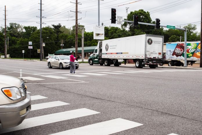 image A Black woman crosses in a crosswalk in a very wide busy road as a semi truck zooms by.