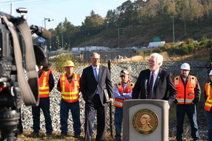 29381999463_8a97c662b3_o Millar wears a suit and stands at a lectern with Governor Inslee and a group of orange vest wearing construction workers standing behind him. A busy freeway is in the background.