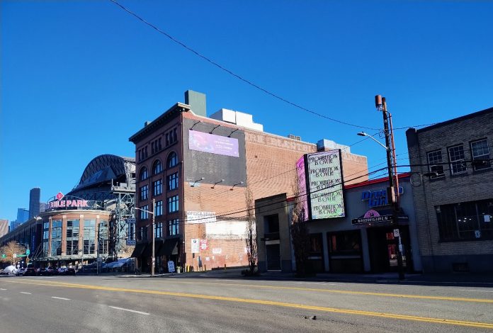 SoDo STOD A photo from SoDo, with T-Mobile Park in the background. An older brick building stands next to smaller, more run down storefronts with a large wide road in the foreground