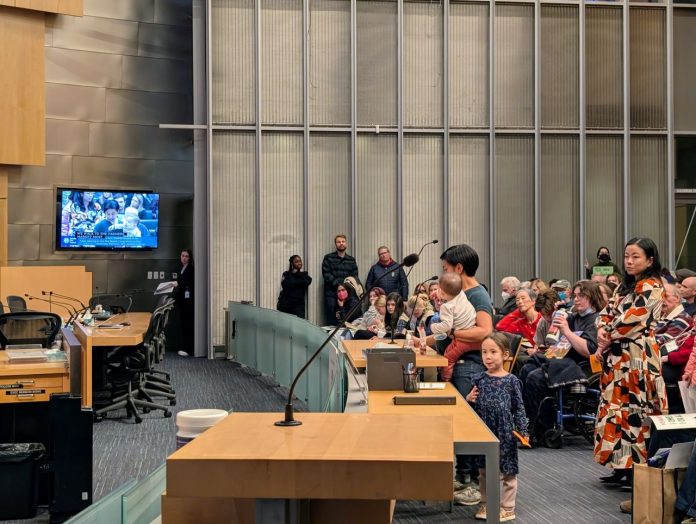 Mother and two kids testifies at One Seattle Hearing_20250206 The Urbanist A women holds a baby with a toddler at her feet and speaks into the micro in Seatlte council chambers.