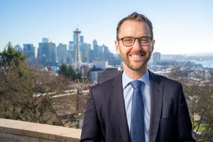 Nathan Rouse Headshot 1600px A white man in glasses and suit poses in from of the Seattle skyline