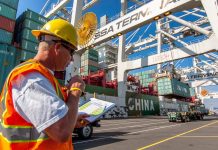 Op-Ed: SoDo Is for Industry, Seattle Should Reject Rushed Housing Push A cargo checker in a yellow hardhat looks at a clipboard with a stack of shipping bins and big crane in the background,