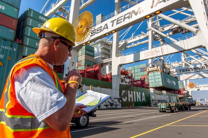 Port of Seattle terminal T30_100929_030 A cargo checker in a yellow hardhat looks at a clipboard with a stack of shipping bins and big crane in the background,