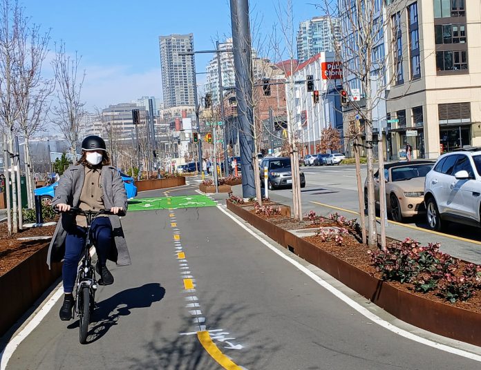 20250301_110945 A person on a Brompton folding bike wearing a mask and biking south on Alaskan Way's bike lane near Seneca Street