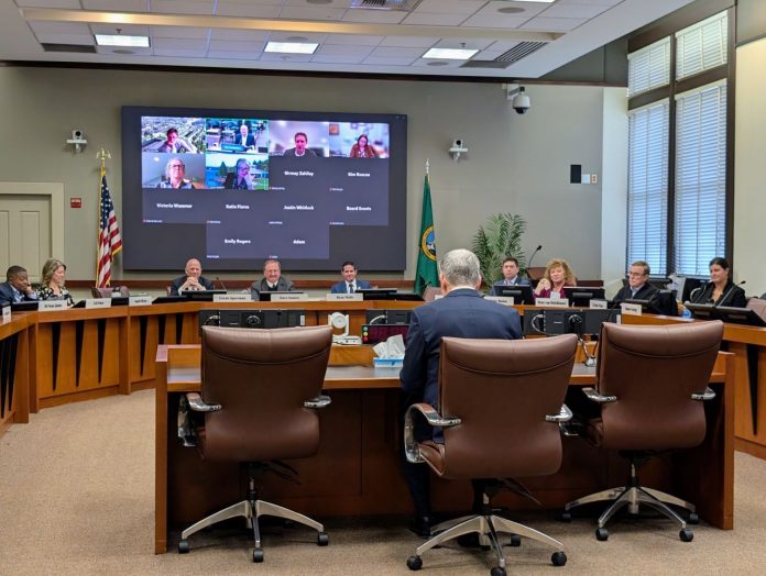 Constantine addresses Sound Transit Board - The Urbanist 20250327 Constantine sits at fishbowl desk with a U-ring of seated boardmembers around him and a video screen with the members joining online.