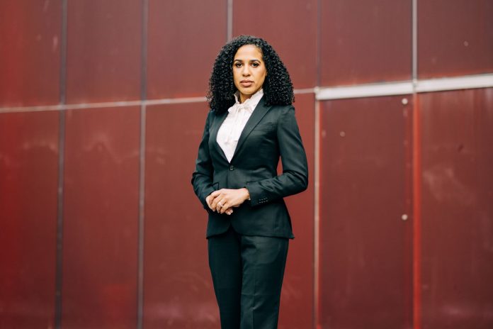 Erika Evans campaign photo 20250326 Evans is a light-skinned Black woman with curly hair standing in front of a red wall.