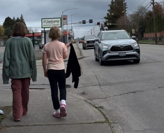 Girls on 6th by Travis Merrigan Two girls walk down 6th narrow sidewalk as cars zip by. One draps a jacket over her shoulder.