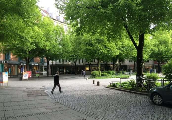 Munich Plaza with Trees - Mike Eliason A quiet pedestrian street with a few people walking and plenty of tree canopy with a midrise residential building in the background.