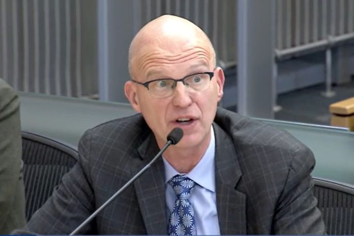 Nathan Torgelson via Seattle Channel Dec 2023 Torgelson wears glasses, a blue checkered tie and a dark suit.