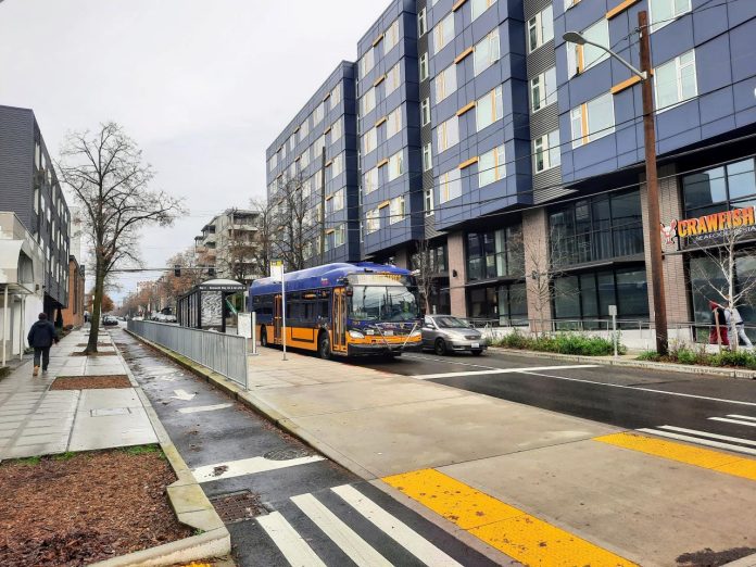 Roosevelt crawfish apartment buildig with bus - Trumm 20231218 A travels down Roosevelt Way next to the protected bike lane. A large seven-story apartment building with a crawfish restaurant on the first floor is on the right.
