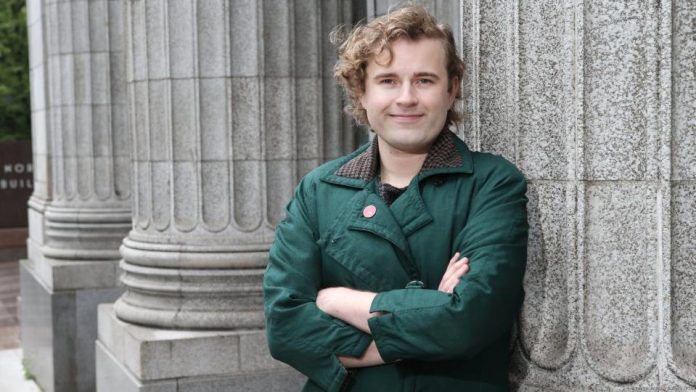 Ry Armstrong campaign photo 2025 Ry stands in front of a Greek column with their arms crossed