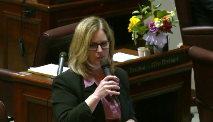 Cleveland hold the microphone on the floor and has blond hair and glasses. On the Annette Cleveland desk behind her is a bouquet of flowers.