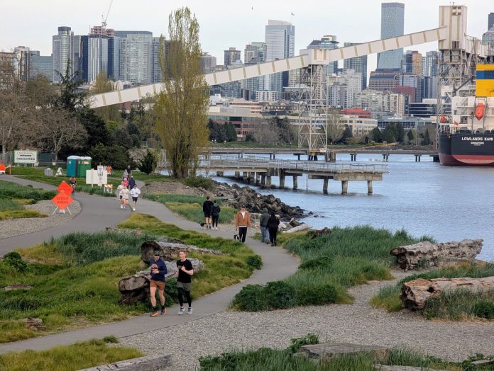Elliott Bay Trail joggers at Expedia Park with skyline - Trumm 20250416 Orange signs warn of the bike path closure just east of Expedia Park. A dozen joggers and dog walkers crowd the waterfront path. The Seattle skyline is in the distance.