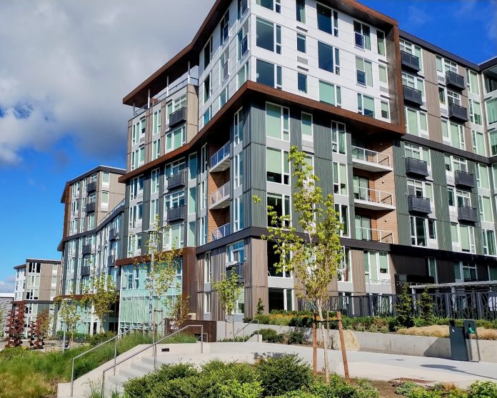 A large midrise building with some landscaping on the edges and blue skies and a fluffy cloud overhead.
