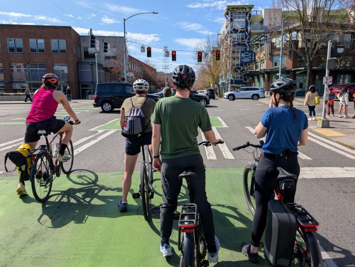 Five people on bike wait in a green bike lane at a major intersection with car whizzing by and pedestrians waiting on the corner.