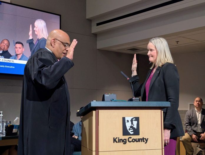 A judge in black robes swears in Braddock who raises her right hand and stands next a lectern with King County's MLK logo. She is a middle-aged blond woman.